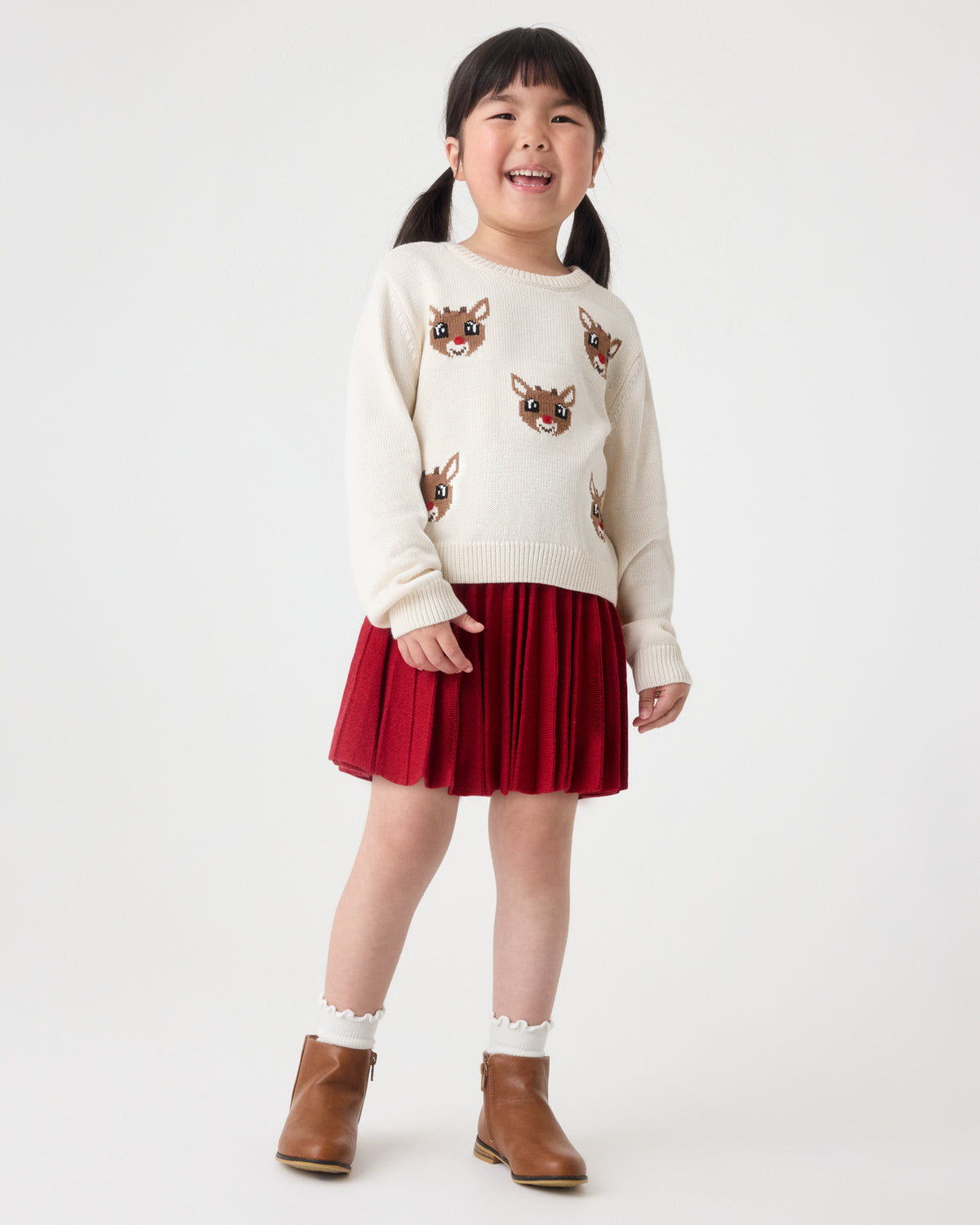A young girl stands against a plain light gray studio background, wearing a cream knit sweater with reindeer faces, a dark red pleated skirt, white socks, and brown ankle boots.