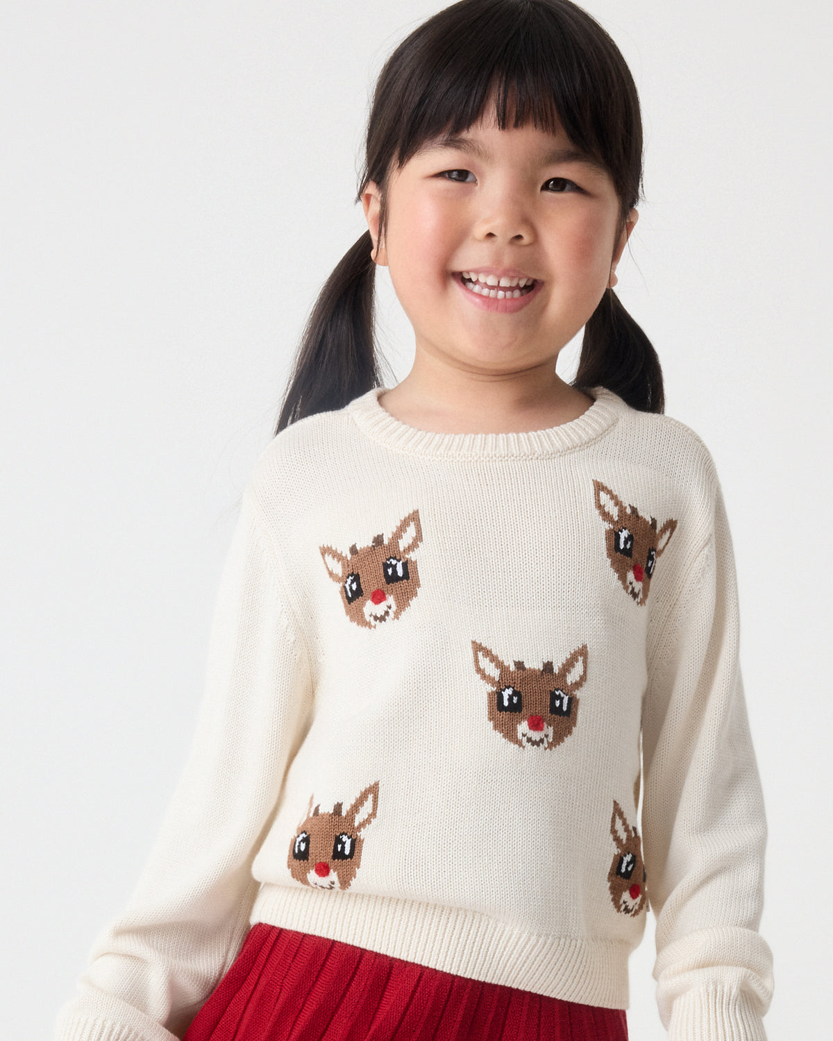 A young girl wearing a cream-colored knit sweater with brown reindeer faces and a red skirt, against a plain light gray background.