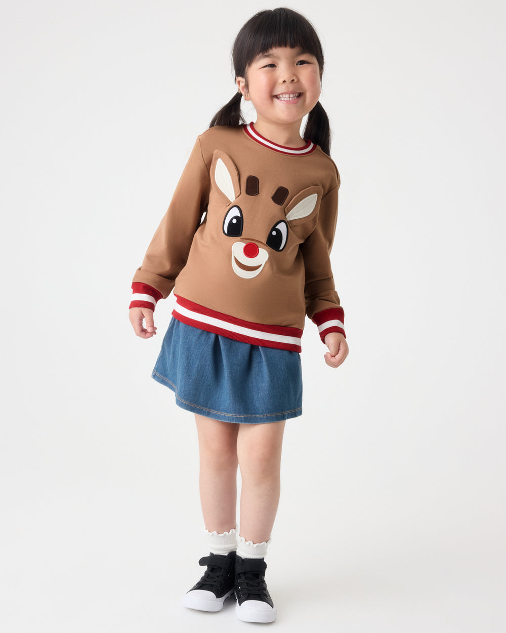 A young girl stands against a light gray studio background wearing a brown long-sleeve sweater with a reindeer face graphic, a blue denim skirt, white socks, and black sneakers.