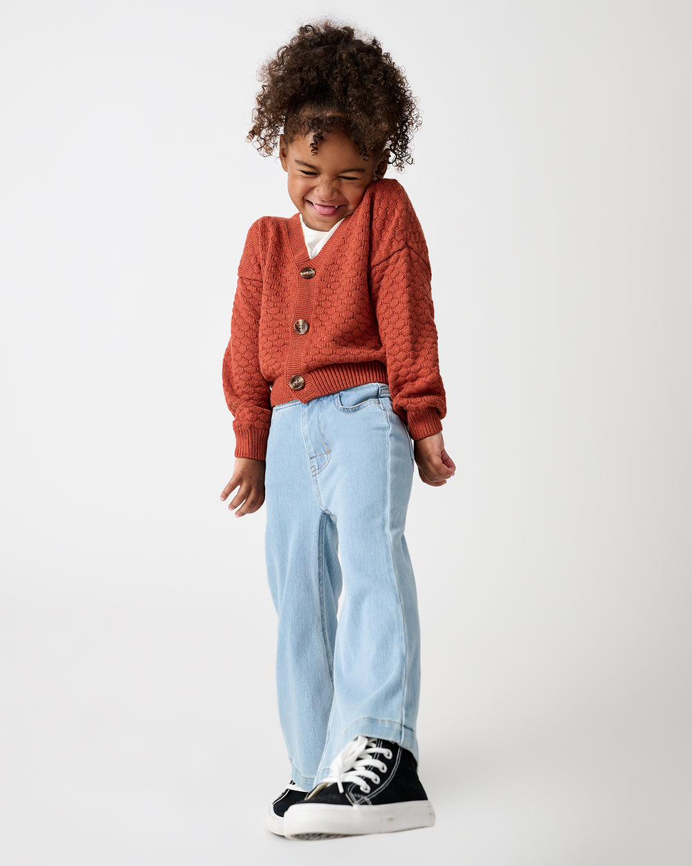 A young child stands against a light gray studio background, wearing a rust-orange textured cardigan over a white shirt, light blue jeans, and black sneakers.