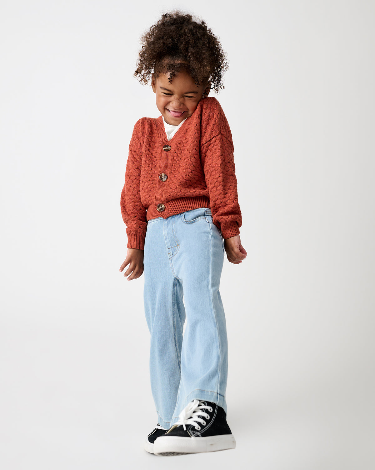 A young child stands against a light gray studio background, wearing a rust-orange textured cardigan over a white shirt, light blue jeans, and black sneakers.