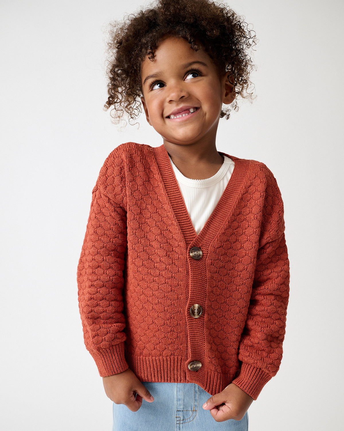 A smiling young child with curly hair wearing a textured rust-orange knit cardigan over a white t-shirt and light blue jeans, photographed against a clean white studio background.