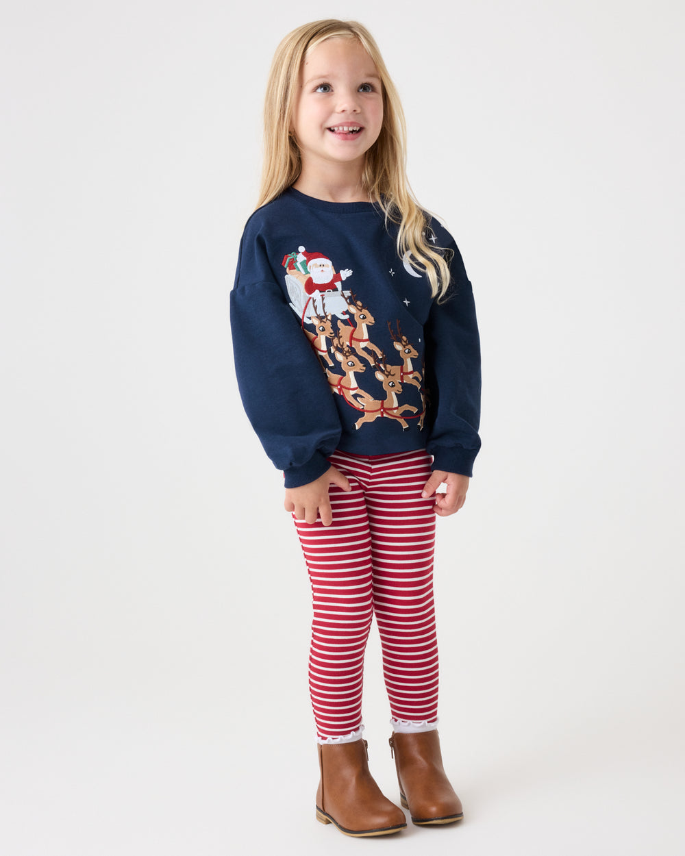 A young girl with blonde hair stands against a white studio background wearing a navy blue crewneck sweatshirt with a Christmas Santa graphic, red and white striped leggings, and brown ankle boots.