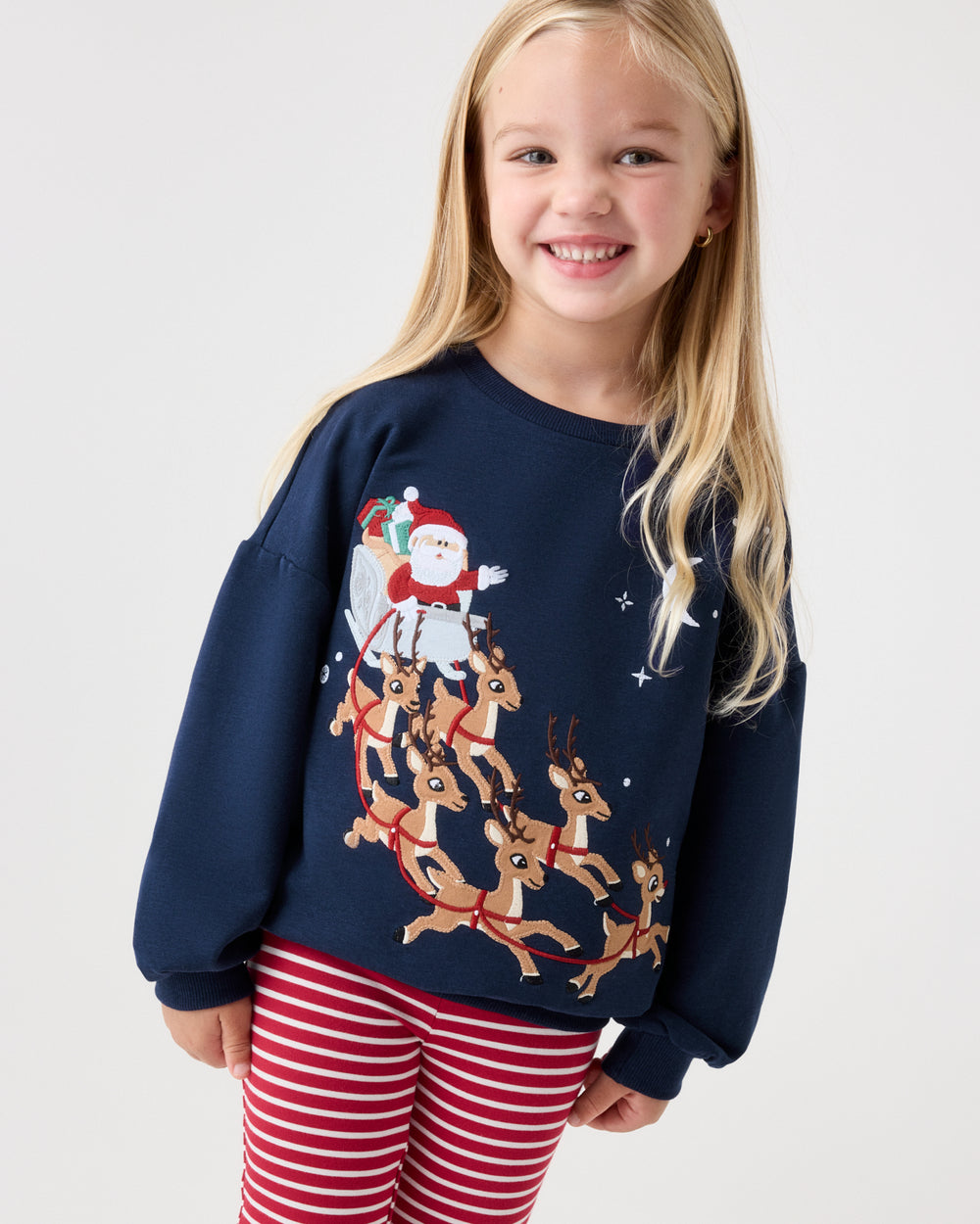 A young girl with blonde hair smiling, wearing a navy blue crewneck sweatshirt with a Christmas graphic of Santa Claus and his reindeer, paired with red and white striped leggings, against a plain light gray background.