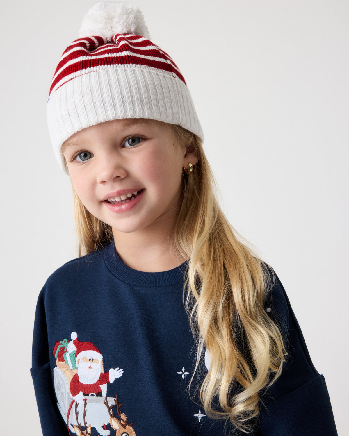 A young girl wearing a white and red striped knit beanie with a pom-pom and a navy blue Christmas sweatshirt featuring a Santa Claus graphic, photographed against a light gray studio background.