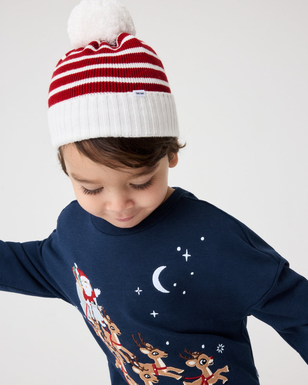 A child wearing a navy long-sleeve Christmas pajama top with a Santa Claus and reindeer graphic, paired with a red and white striped knit beanie with a white pom-pom, against a light gray studio background.