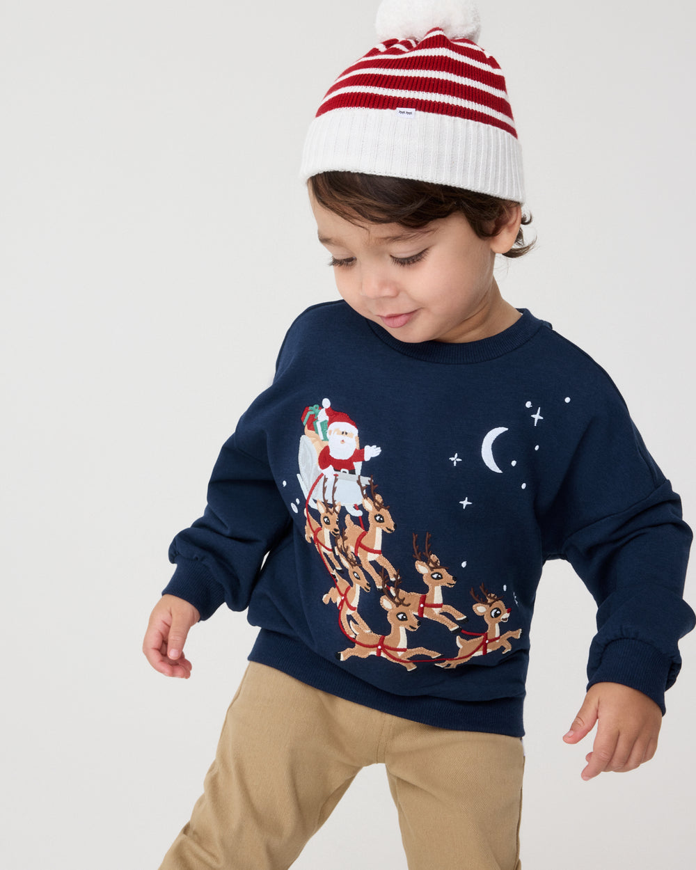 A toddler wearing a navy blue Christmas crewneck sweatshirt with a Santa Claus and reindeer graphic, paired with beige pants and a red and white striped knit beanie, against a light gray studio background.