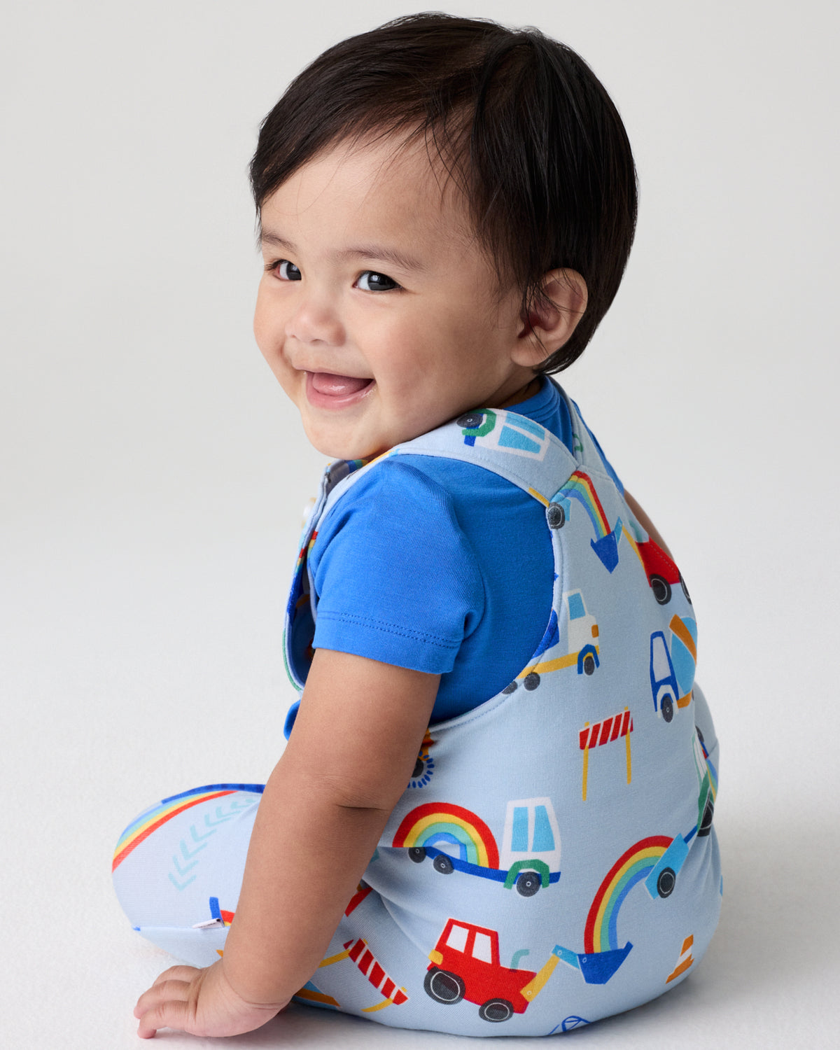 Back view of sitting baby wearing the Rainbow Trucks Overall Set