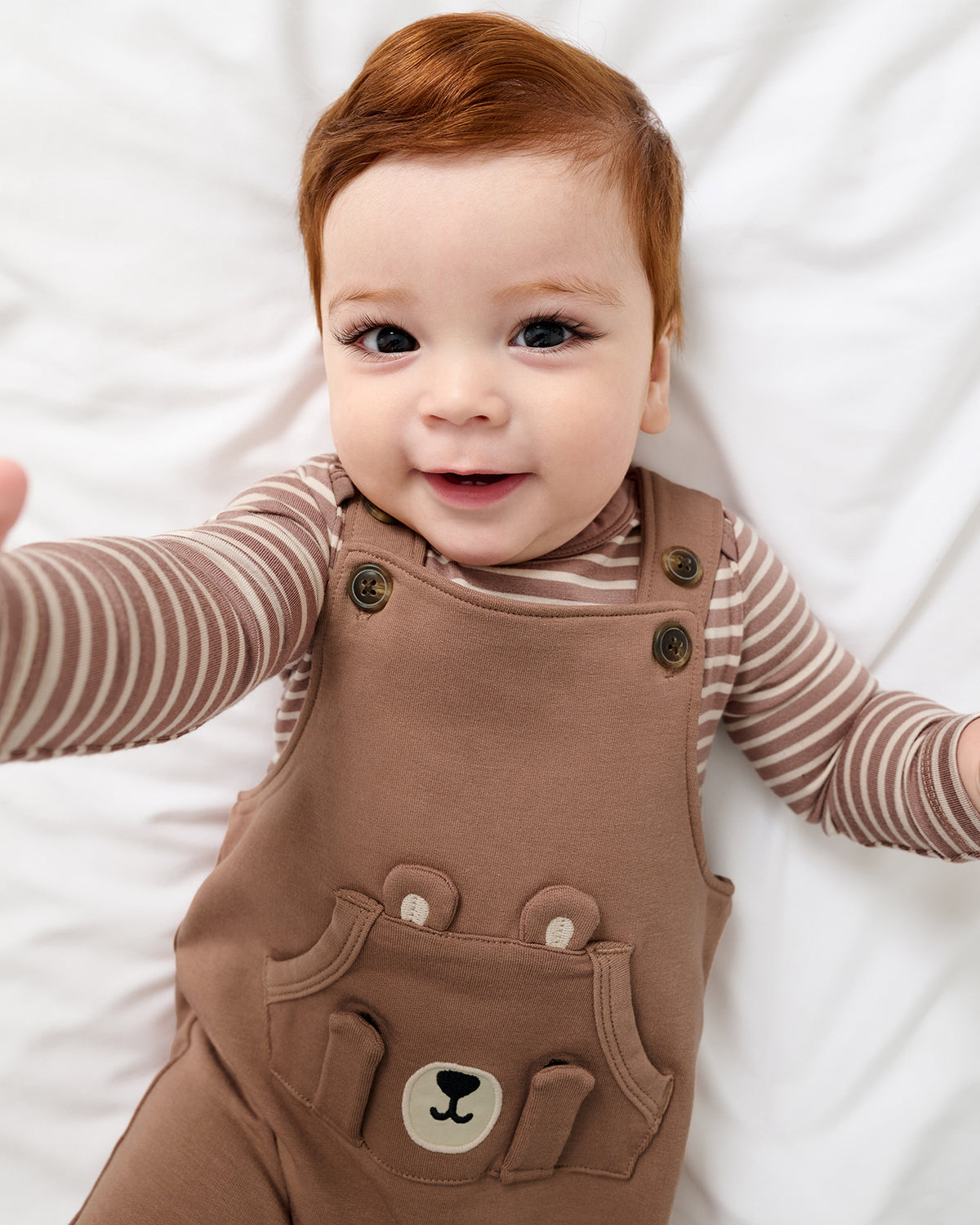 A smiling baby with red hair lies on white bedding, wearing brown overalls with a bear face patch pocket over a beige and brown striped long-sleeve bodysuit.