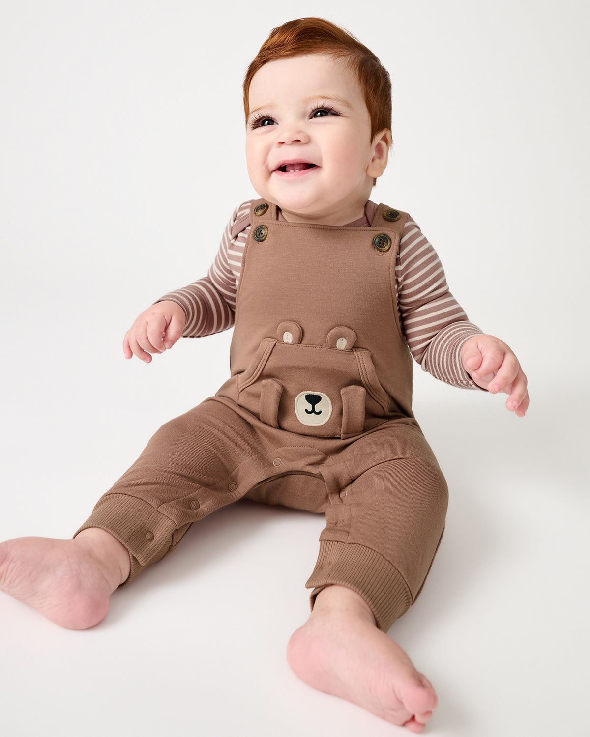A smiling baby with red hair wearing a brown overall romper over a brown and beige striped long-sleeve bodusuit, sitting against a plain white studio background.