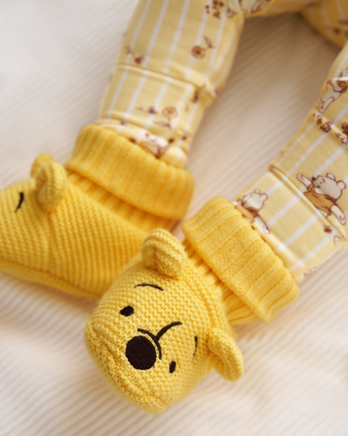 Yellow knit booties with Winnie the Pooh character faces, attached to striped yellow leggings with a bear print, shown on a soft cream-colored fabric background.