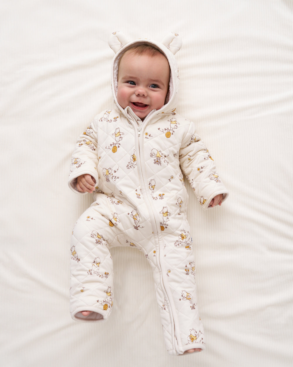 A baby lying on a white textured blanket, wearing a quilted cream-colored footed snowsuit with a printed pattern of small yellow and brown animals and a hood with bunny ears.