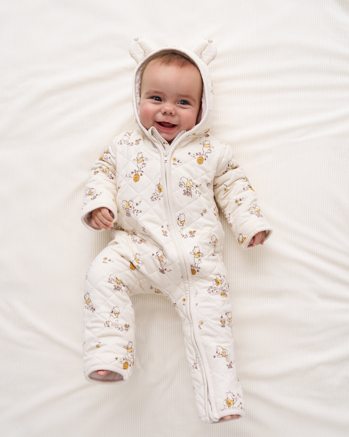 A baby lying on a white textured blanket, wearing a quilted cream-colored footed snowsuit with a printed pattern of small yellow and brown animals and a hood with bunny ears.