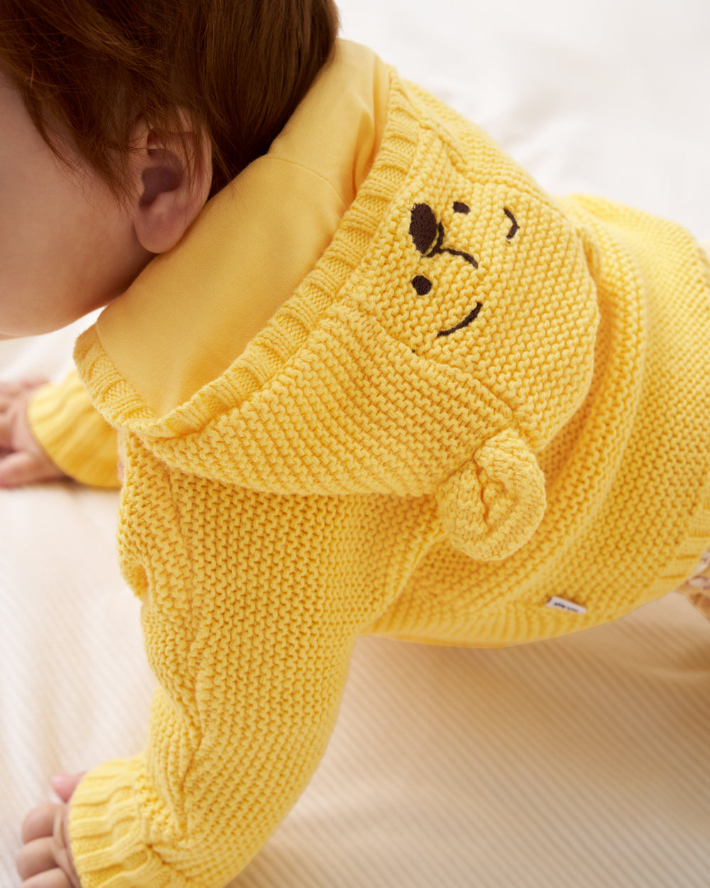 A close-up of a baby wearing a bright yellow knitted hooded sweater with a Winnie the Pooh-style embroidered face on the hood, crawling on a light cream fabric.