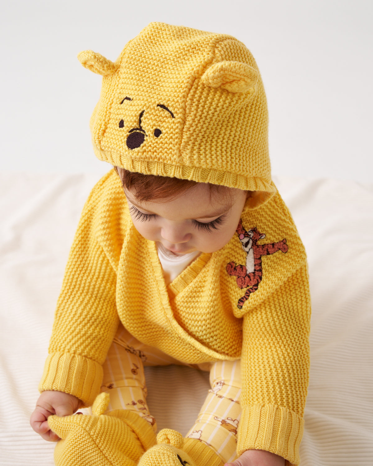 A baby wearing a yellow knit hooded cardigan and matching booties, styled like Winnie the Pooh. The hood features embroidered faces of Pooh and Tigger, and the cardigan has an embroidered Tigger design on the chest.