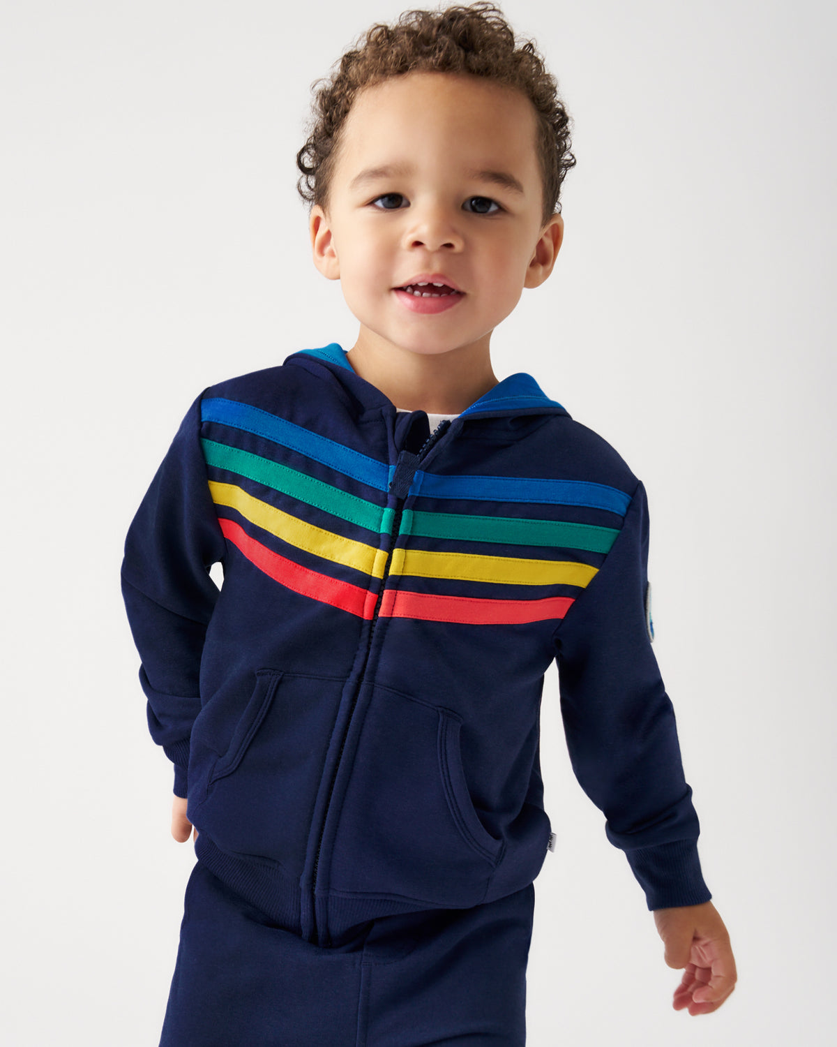 A toddler wearing a navy blue zip-up hoodie with colorful horizontal stripes across the chest in blue, green, yellow, and red, paired with matching navy pants, photographed against a light gray studio background.