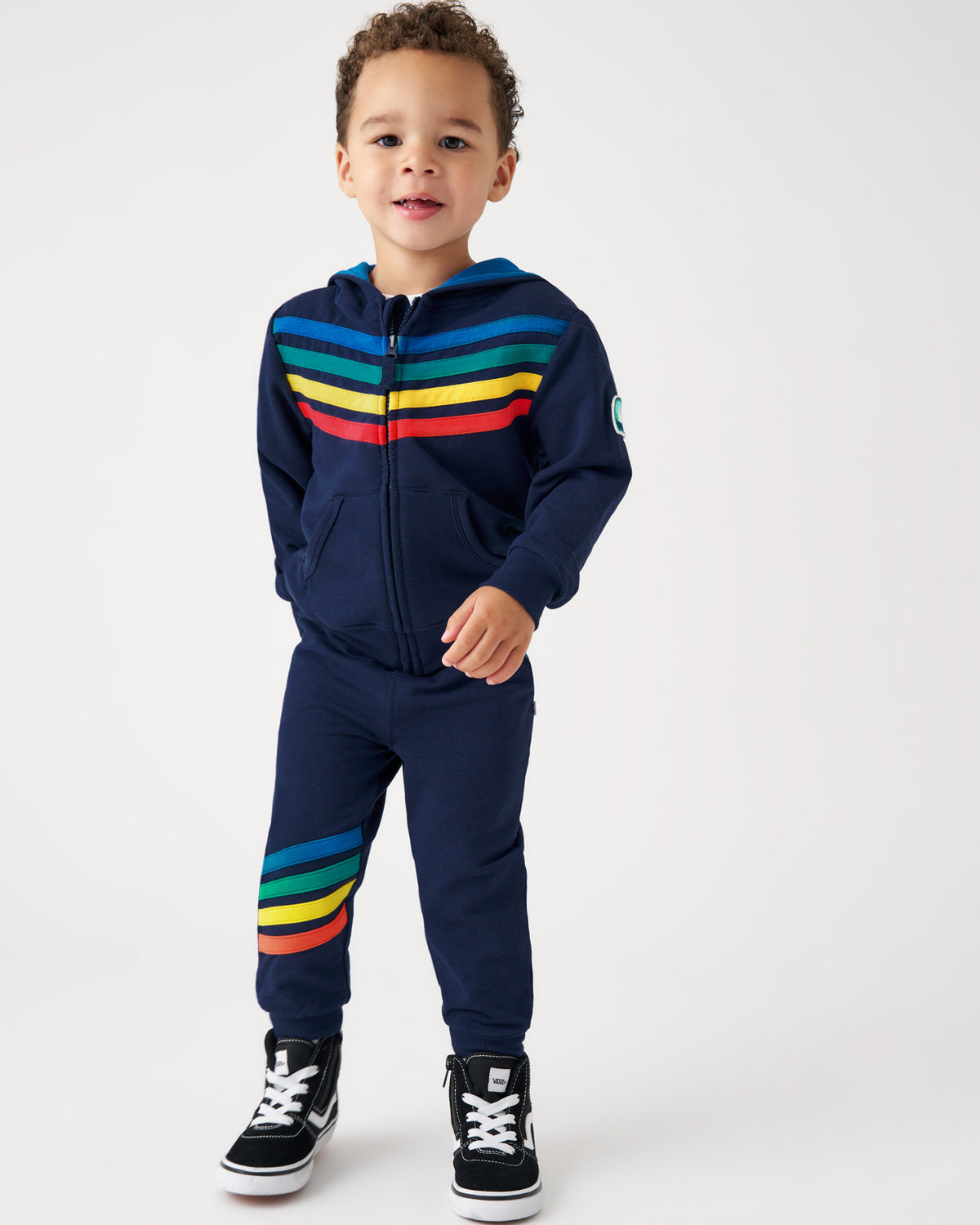 A toddler boy wearing a navy blue zip-up hoodie and matching sweatpants with rainbow stripes, paired with black sneakers, standing against a light gray studio background.