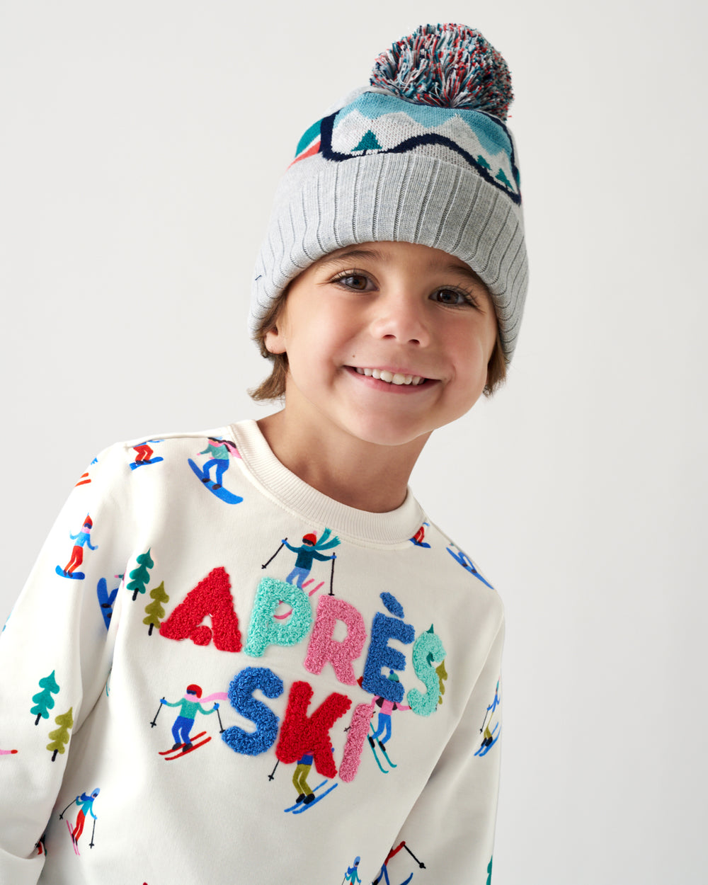 A smiling young boy wearing a gray knit pom-pom beanie and a cream sweatshirt with colorful ski-themed lettering and illustrations, photographed against a clean light gray studio background.