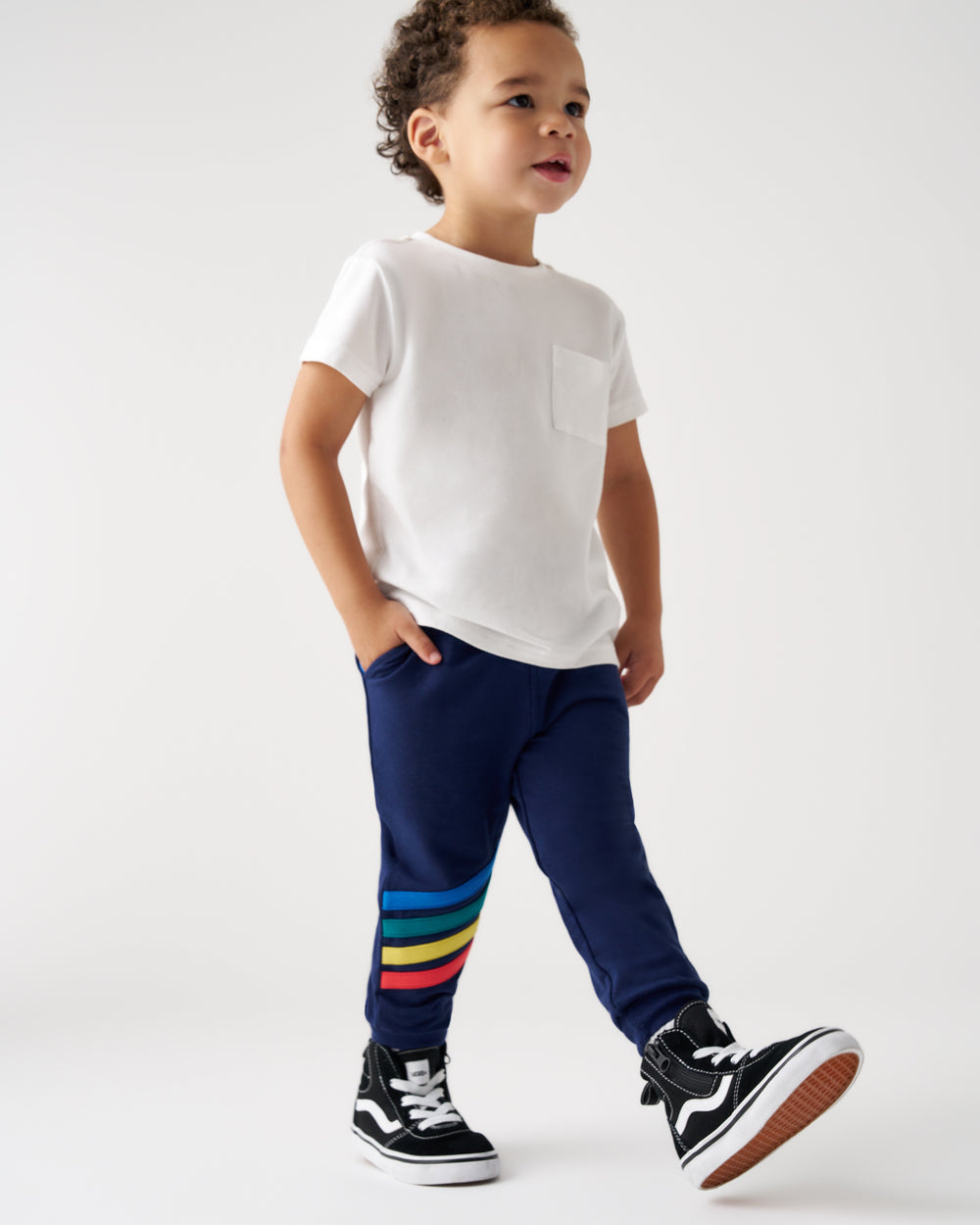 A toddler standing in a studio wearing a white short-sleeve pocket T-shirt, navy pants with a colorful striped detail near the lower leg, and black-and-white sneakers.