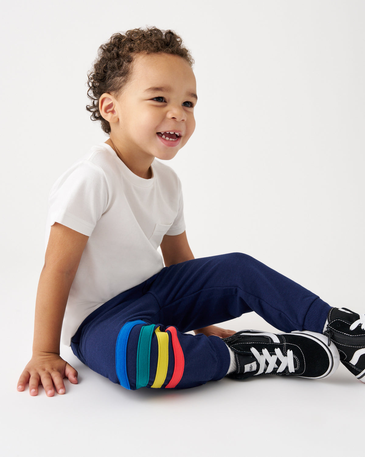 A smiling young boy with curly hair sits on a white studio floor wearing a white t-shirt, navy pants with colorful elastic leg bands, and black sneakers.