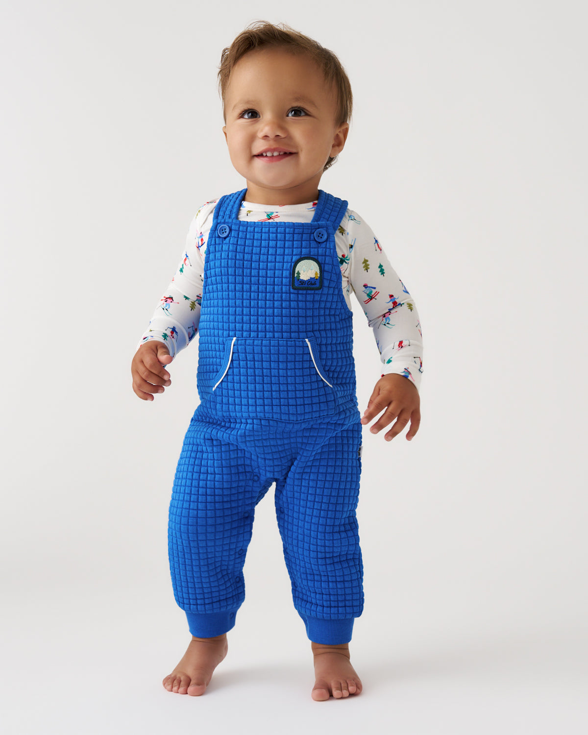 A smiling toddler standing barefoot against a plain light gray studio background, wearing bright blue grid-patterned overalls over a white long-sleeve shirt with small multicolor cartoon prints.