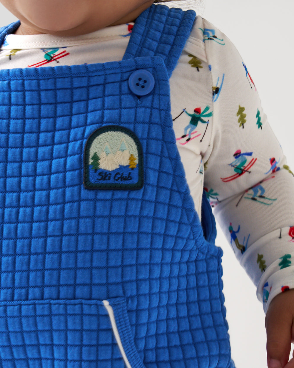 A close-up of a child wearing a bright blue quilted bib overalls with a 'Ski Club' embroidered patch on the chest, layered over a white long-sleeve shirt with a colorful ski-themed print.