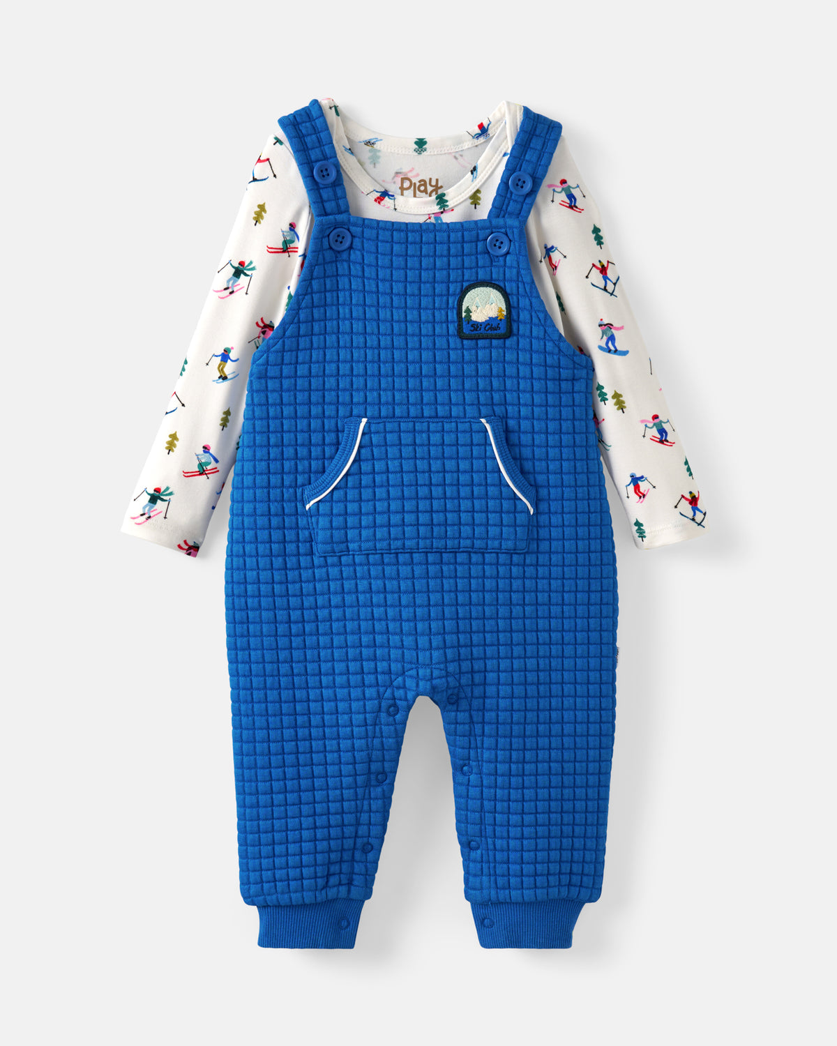 A baby's blue quilted overall jumpsuit with a white long-sleeve shirt underneath featuring a colorful ski pattern. The jumpsuit has a small patch on the chest pocket and is shown against a light gray background.
