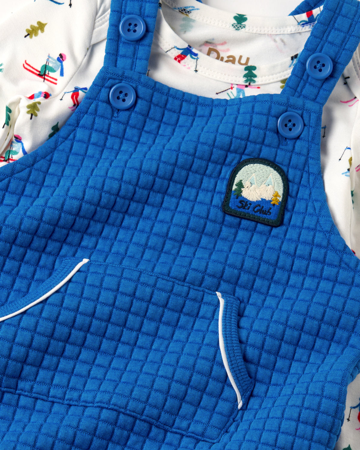 A close-up of a baby's quilted blue overalls featuring a 'St. Cloud' embroidered patch on the chest, layered over a white long-sleeve shirt with a colorful ski-themed print.