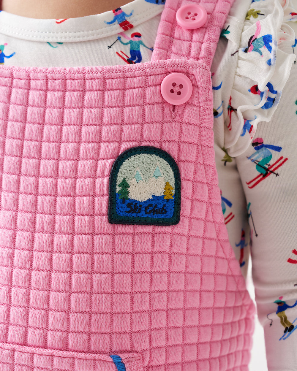 A close-up of a child wearing pink quilted corduroy overalls over a white long-sleeve shirt with a colorful skier pattern. The overalls feature a pink button, a matching pocket, and a small embroidered 'Ski Club' patch on the chest.
