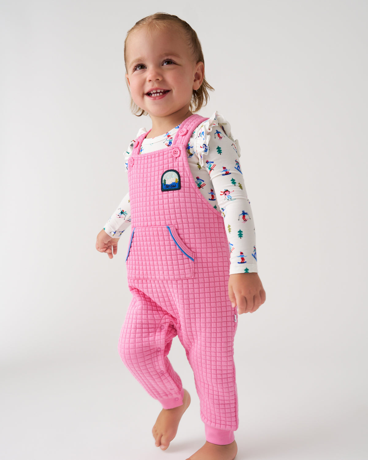 A toddler standing against a light gray studio background, wearing pink grid-patterned overall pajamas over a white long-sleeve shirt with a colorful winter print.