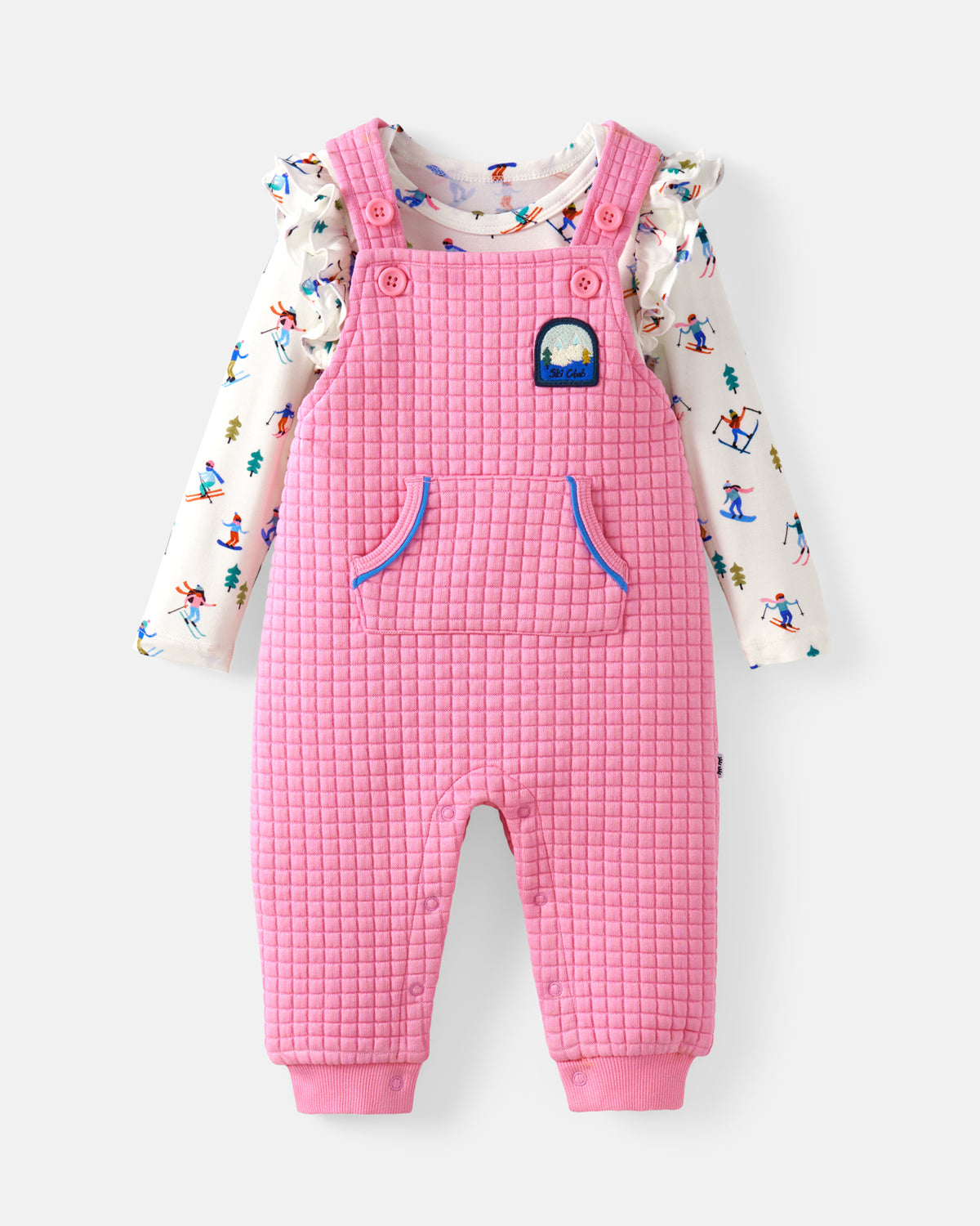 A baby girl's two-piece outfit featuring pink quilted overall jumpsuit and a white long-sleeve ruffle-sleeve shirt with a colorful winter sports print, displayed on a light gray background.
