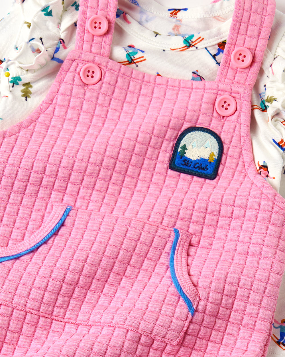 Pink quilted baby overalls with a textured diamond pattern, featuring pink buttons, front pockets with blue piping, and a 'St. Lucia' patch, laid flat on a white patterned fabric background.
