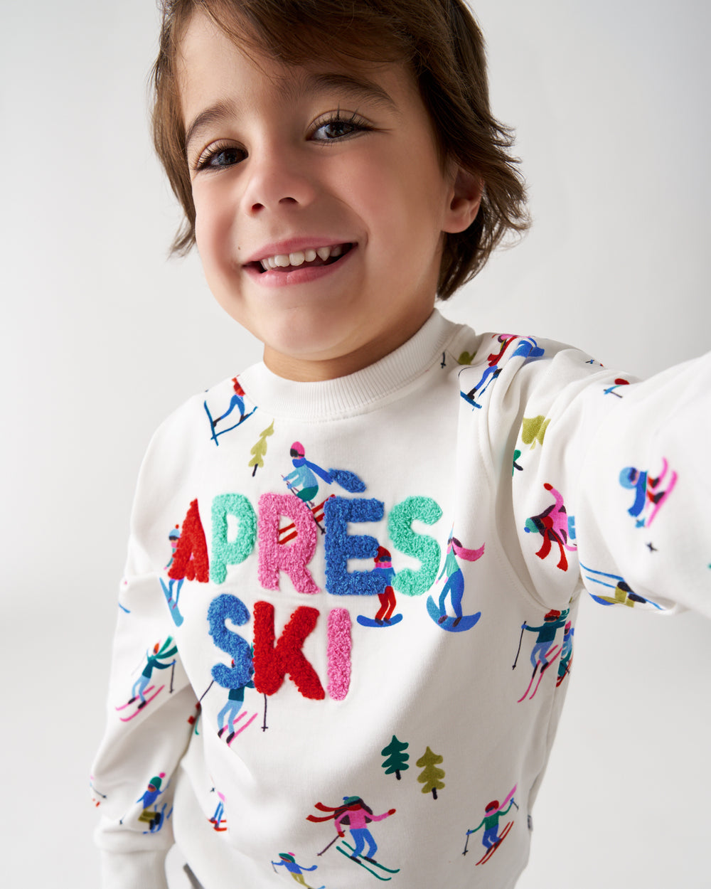 A smiling young boy wearing a white long-sleeve sweatshirt with colorful embroidered text reading 'APRES SKI' and small multicolored skier motifs, photographed against a clean light gray background.
