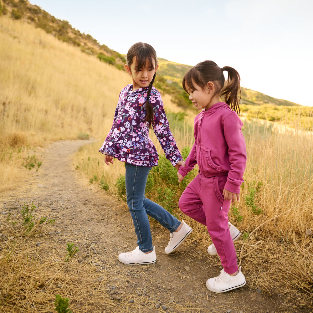 Two girls outdoors walking on a hiking trail. Girl on the right is wearing a Berry Rose Ruffle Hoodie and Jogger. Girl on the left is wearing a Violet Meadow Peplum Hoodie and Midwash Blue Denim Jegging.