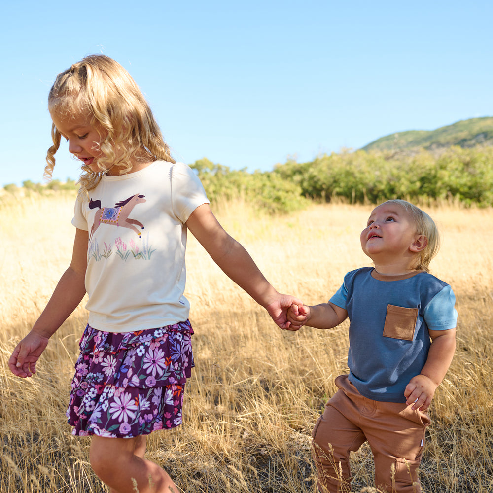 Girl on the left is wearing the Wildflower Ride Puff Sleeve Tee and Violet Meadow Skort and holding boys hand, whose in the Vintage Navy Colorblock Relaxed Pocket Tee and Caramel Cargo Jogger