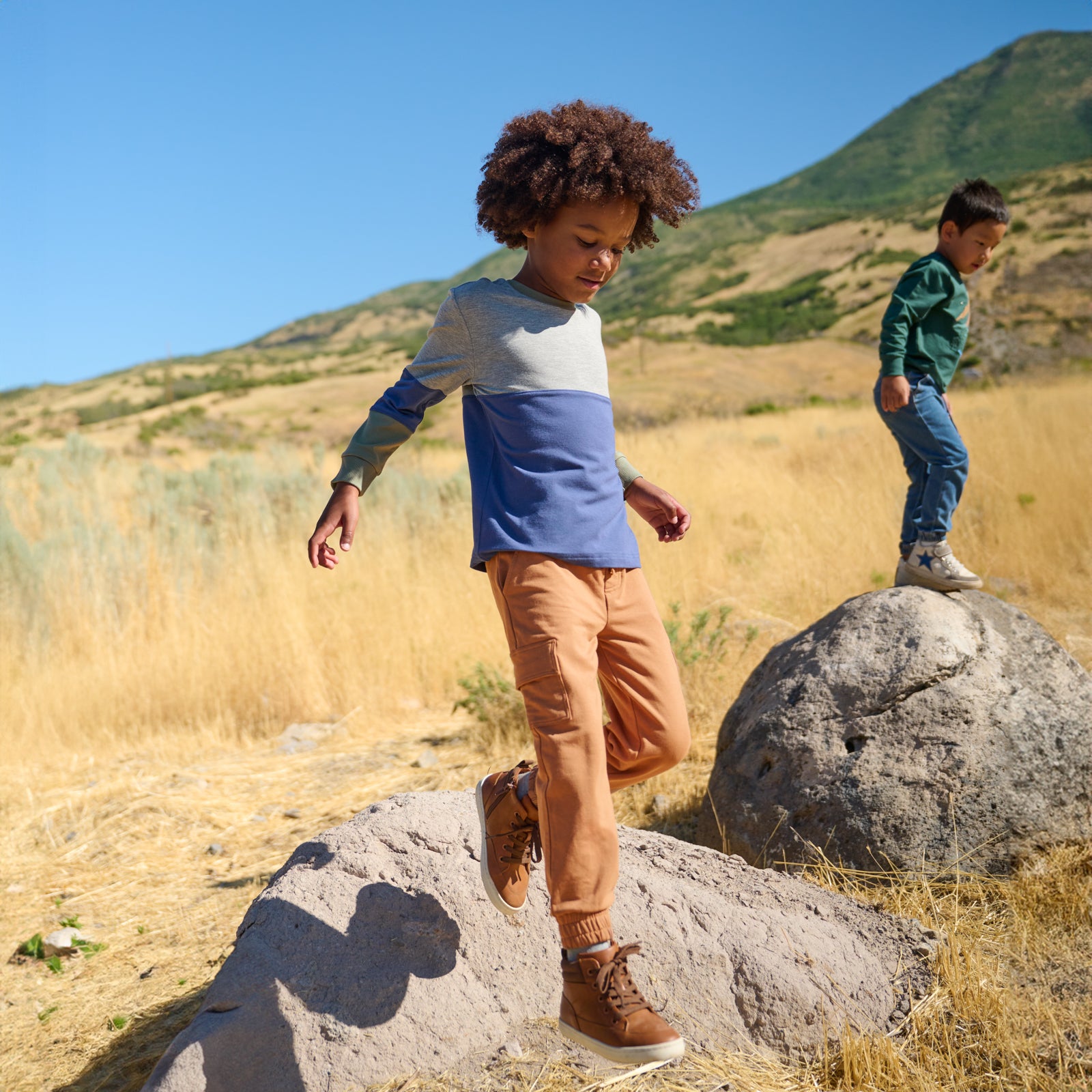 Two boys outdoors climbing rocks. The boy in the front frame is wearing the Vintage Navy Colorblock Relaxed Panel Tee and Caramel Cargo Jogger