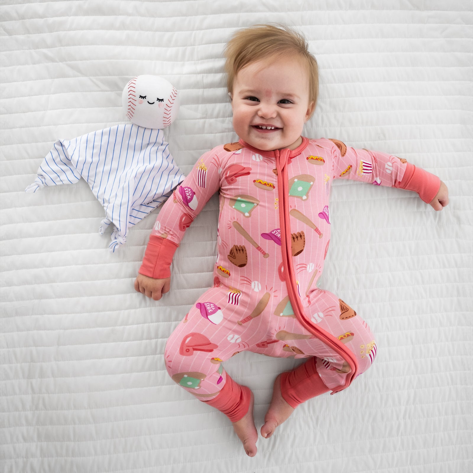 Child laying on a bed wearing Pink All Stars zippy with Major the Baseball sleepyhead lovey
