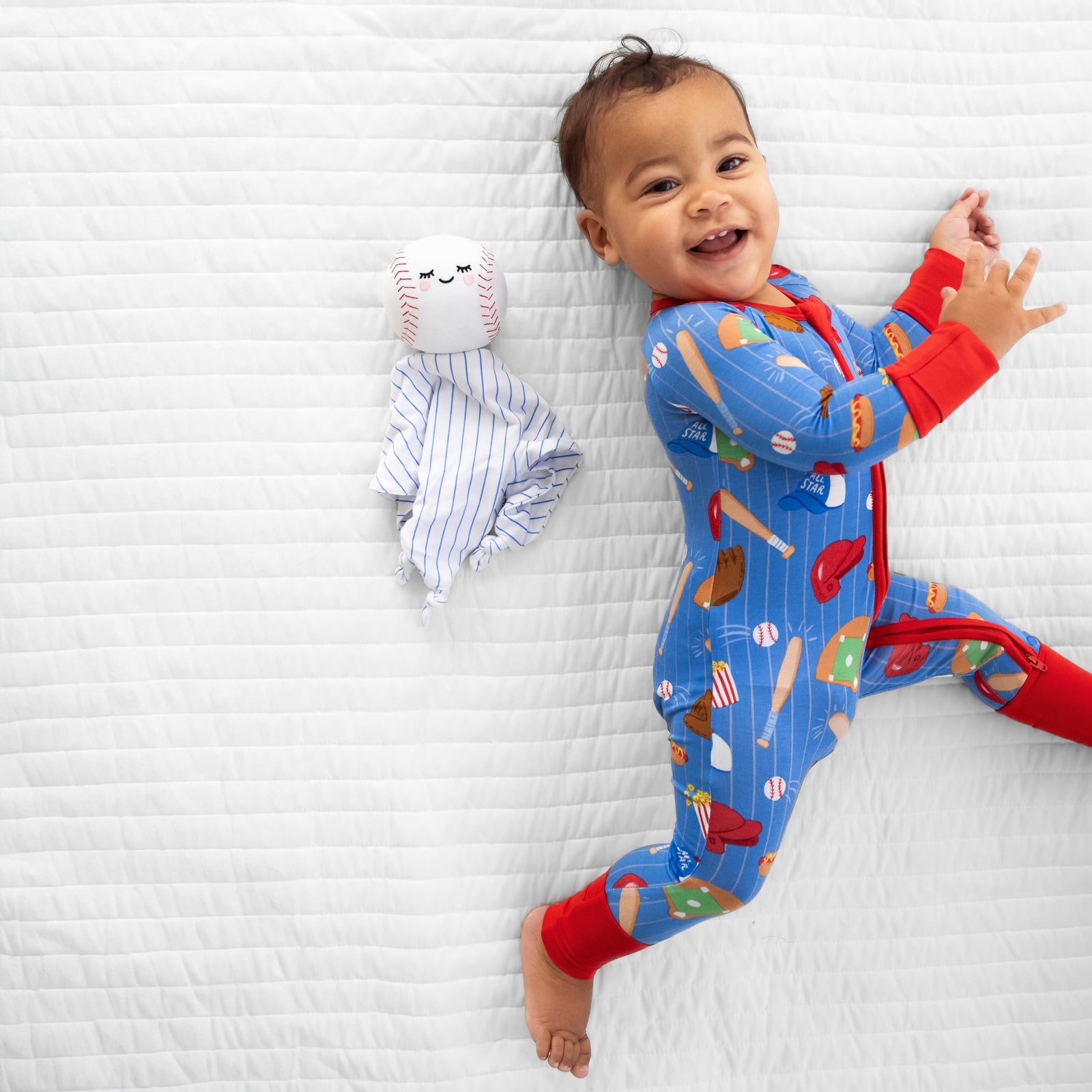 Alternate image of a child laying on a bed wearing Blue All Stars zippy with Major the Baseball sleepyhead lovey
