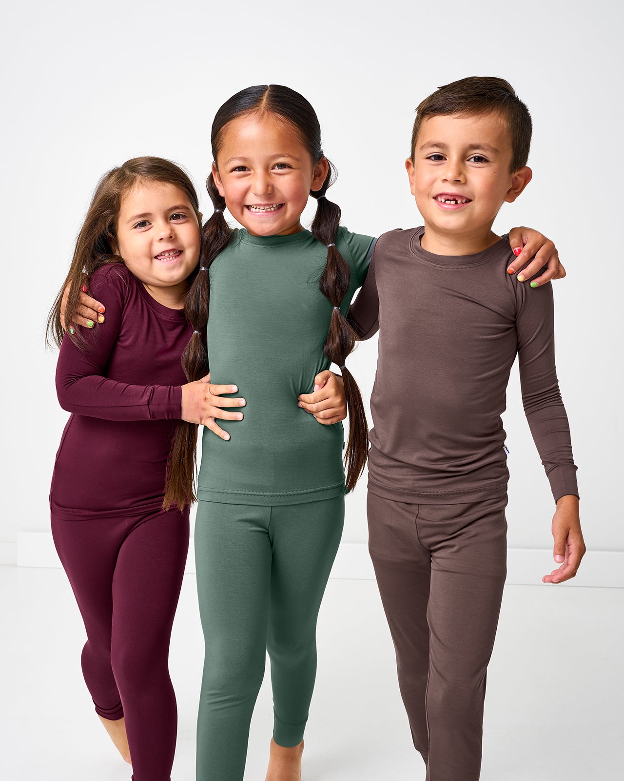 Three children wearing matching solid burgundy, green and brown pajama sets