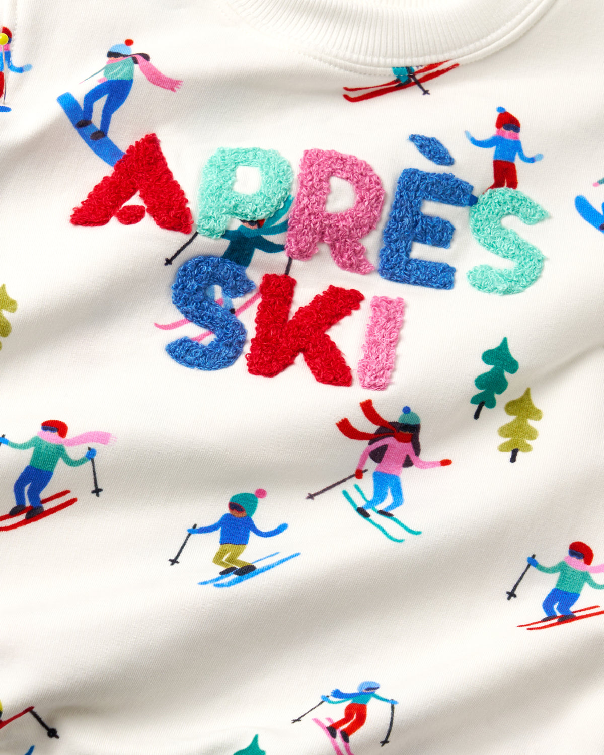 Close-up of a white fleece sweatshirt featuring the word 'APRÈS' in large textured letters, layered with 'SKI' in smaller blue and pink letters. The design is decorated with colorful embroidered ski and snowboard motifs throughout.