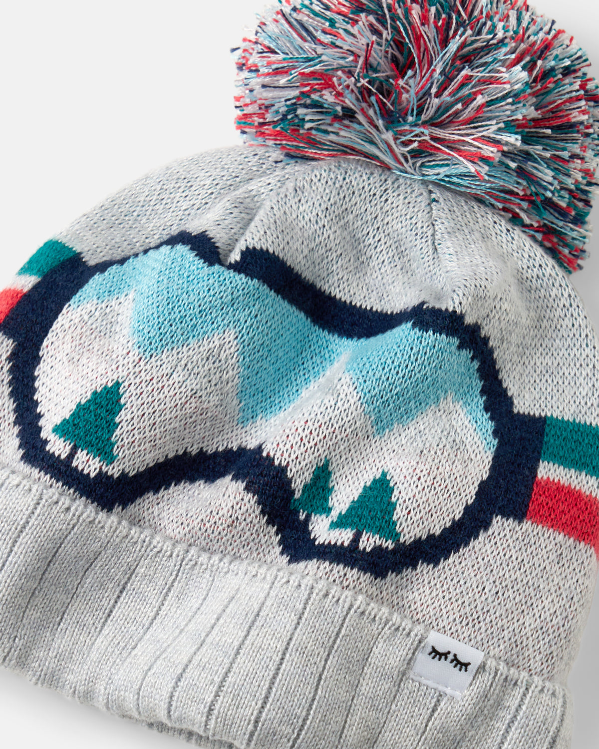 A close-up of a knit pom-pom beanie in light gray, featuring a large ski goggles graphic with a mountain and tree motif in light blue, white, and teal, topped with a multicolor pom-pom.