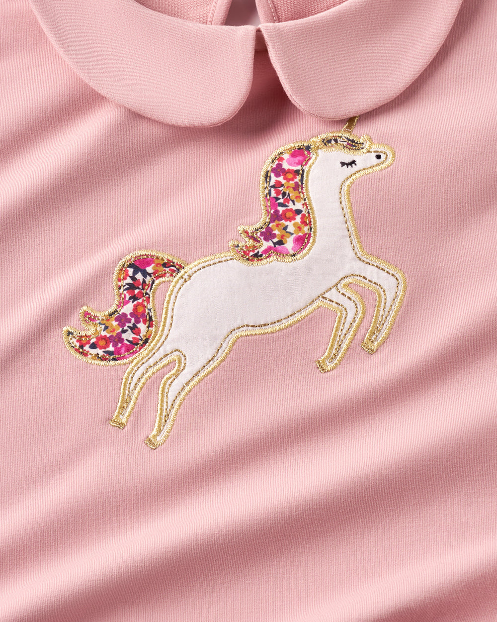 A close-up of a light pink garment featuring an embroidered unicorn applique with a white body, multicolor floral mane and tail, and gold embroidered outlines, set against a matching pink collar.