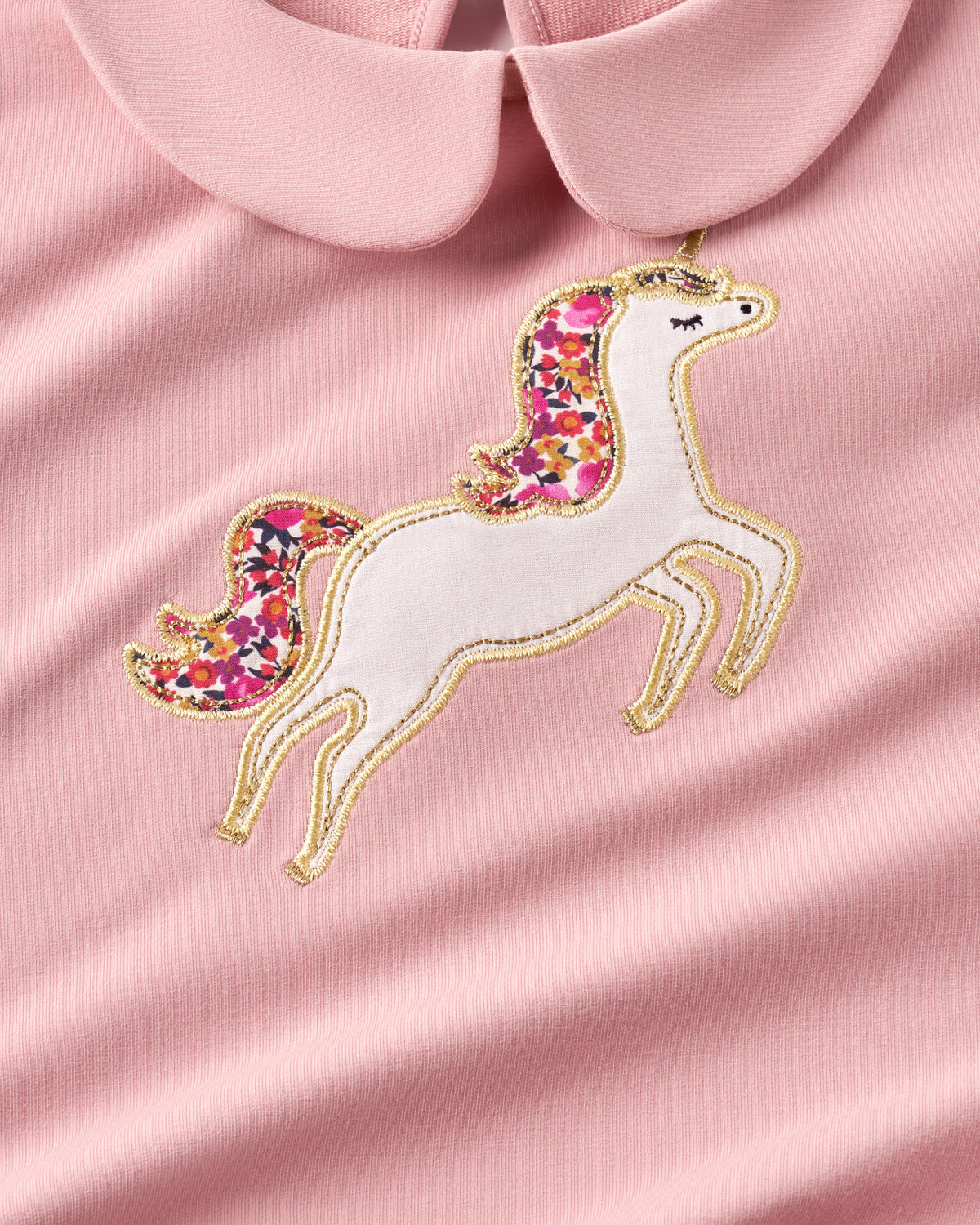 A close-up of a light pink garment featuring an embroidered unicorn applique with a white body, multicolor floral mane and tail, and gold embroidered outlines, set against a matching pink collar.