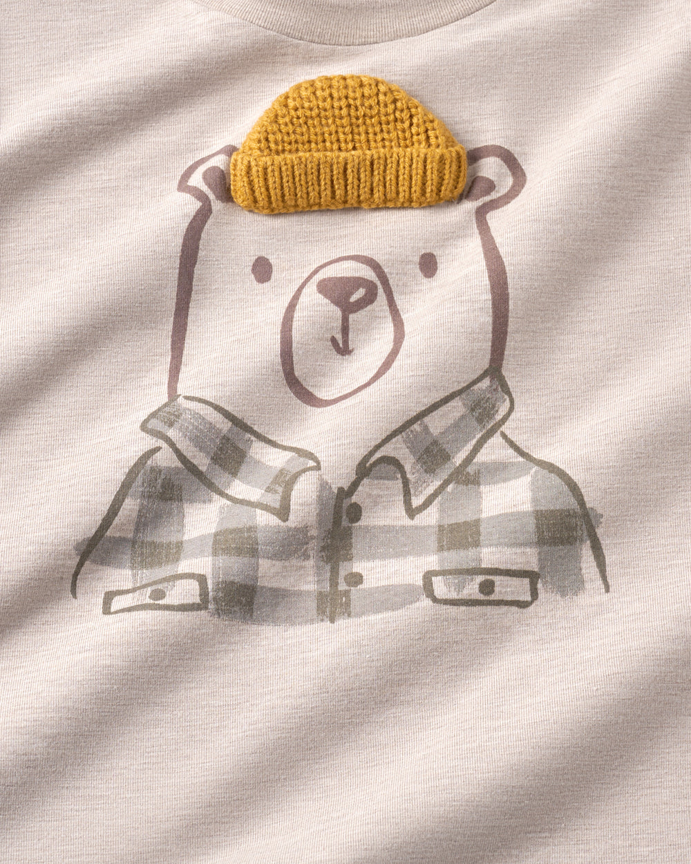 Close-up of a cream-colored t-shirt featuring a cartoon bear graphic wearing a mustard yellow knit beanie and a gray plaid flannel shirt.