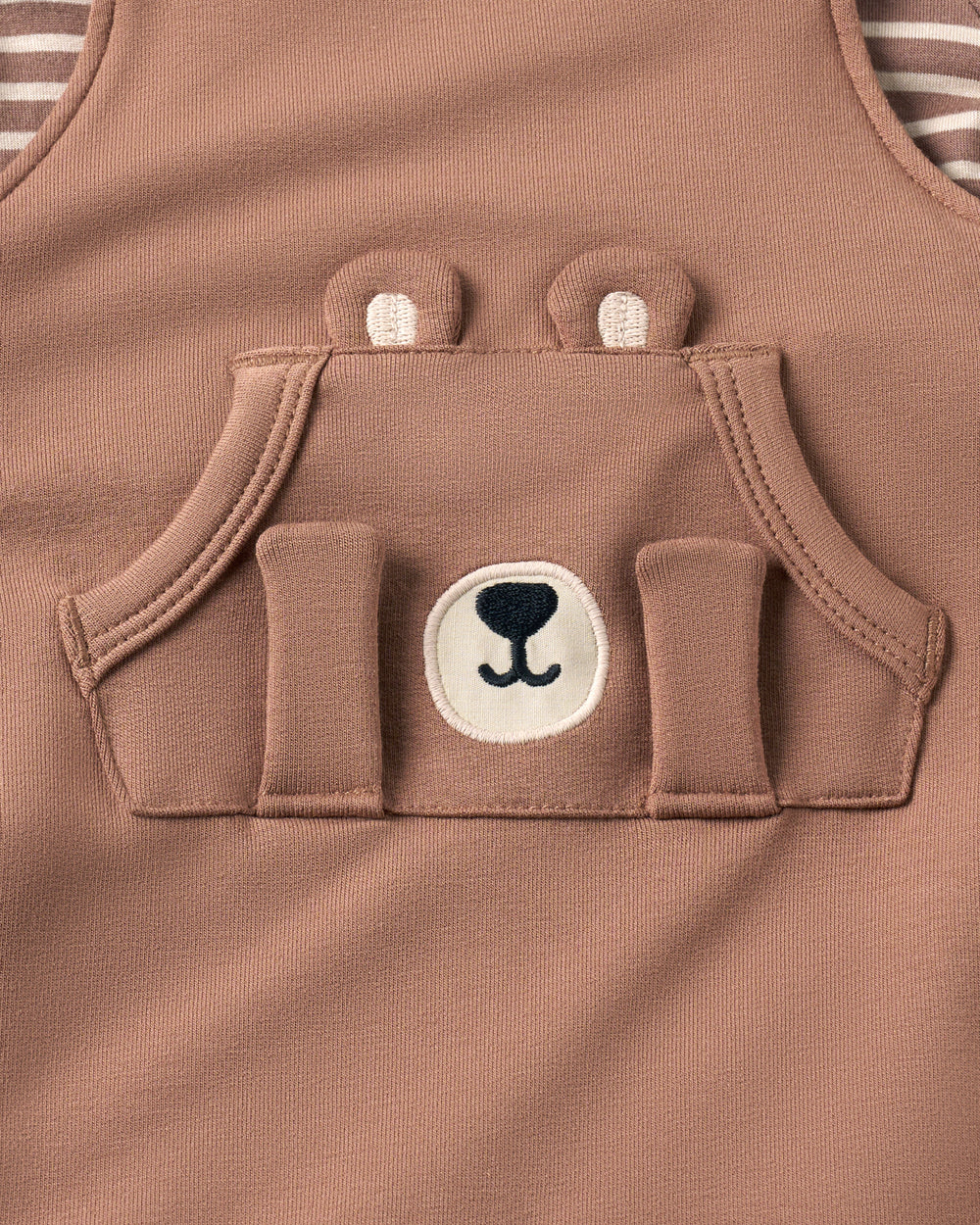 Close-up of a baby romper in a teddy brown color, featuring a front pocket designed to look like a bear face with embroidered eyes, nose, and a striped undershirt visible at the top.