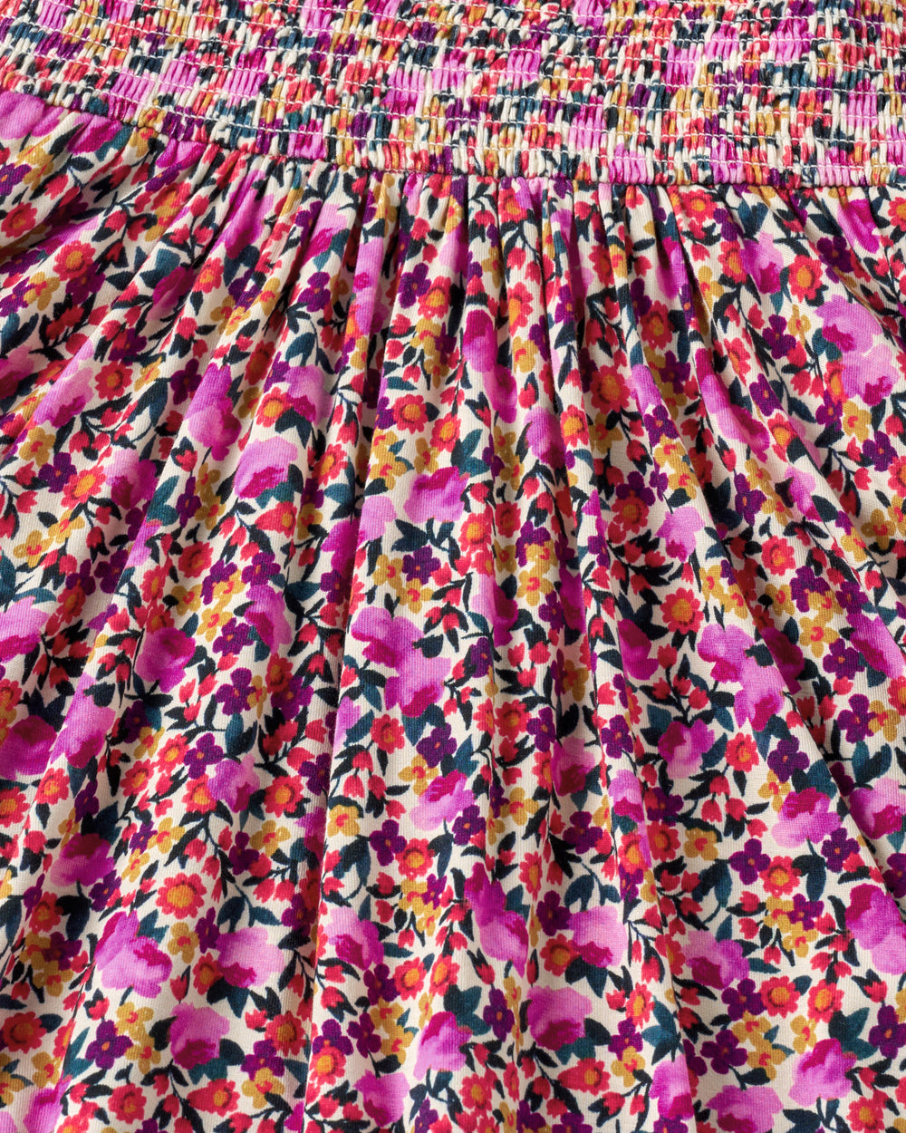 Close-up of a floral skirt fabric with a smocked elastic waistband. The print features pink, purple, orange, and yellow flowers with green leaves on a cream background.
