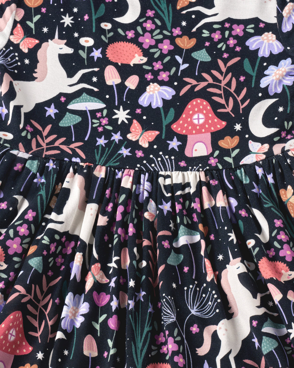 Close-up of a baby dress skirt with a whimsical woodland print on a black background, featuring white unicorns, pink and red mushrooms, purple flowers, green leaves, stars, and crescent moons.