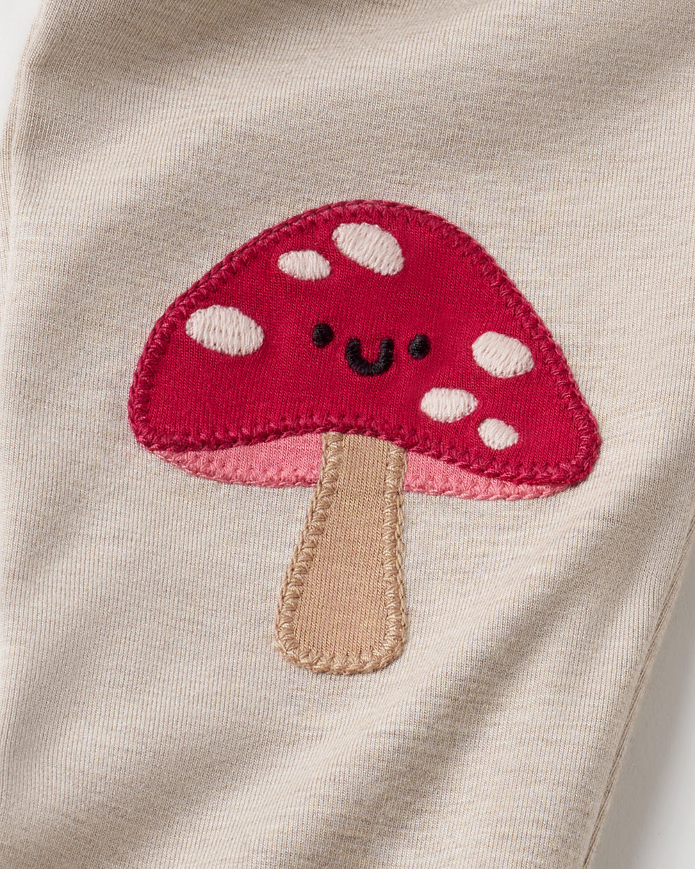 Close-up of a cream-colored knit garment featuring a red embroidered mushroom applique with white spots, a pink underside, and a beige stem.