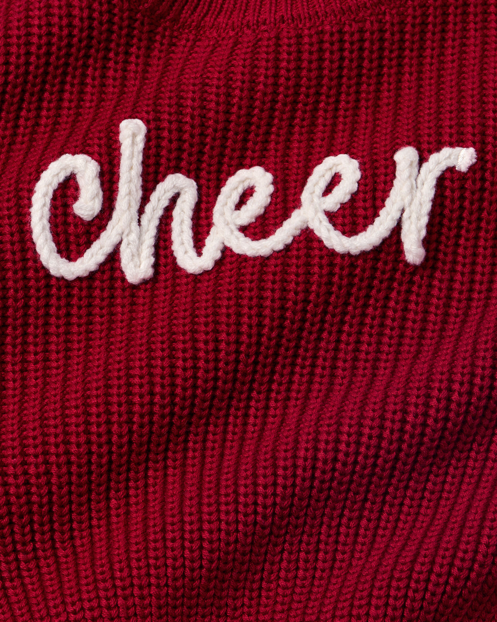 A close-up of a dark red ribbed knit sweater featuring the word 'Cheer' embroidered in white cursive script across the chest.