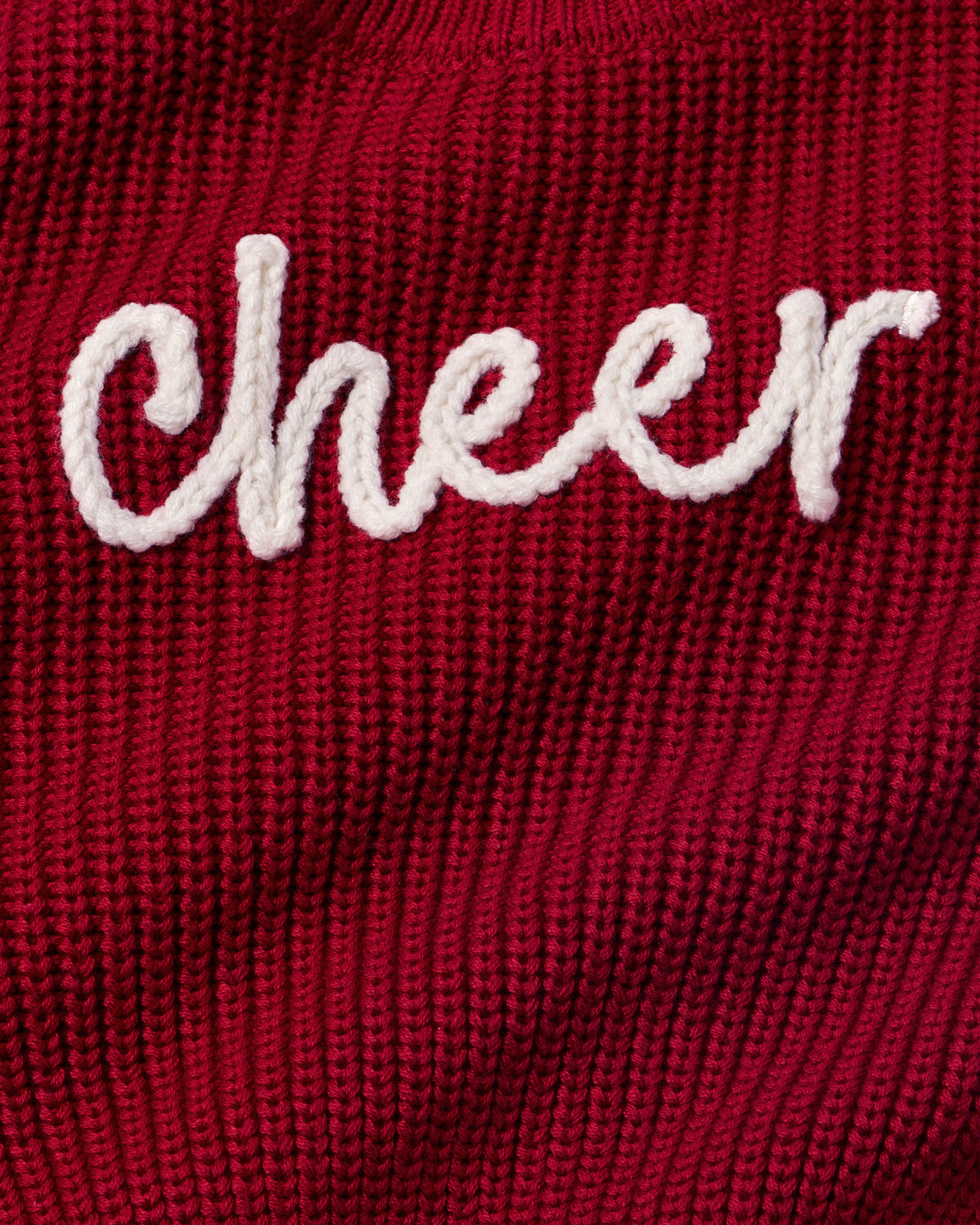 A close-up of a dark red ribbed knit sweater featuring the word 'Cheer' embroidered in white cursive script across the chest.